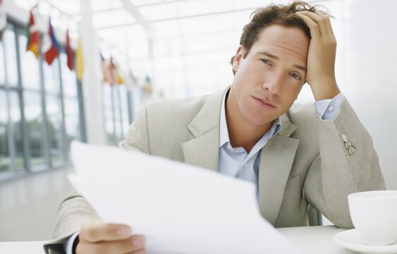 Young male diplomat with brown hair examines tariff documents at a UN desk with a cup of coffee nearby, showing signs of stress