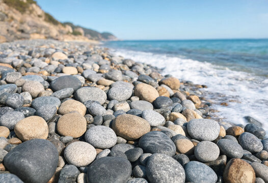 Smooth rounded pebbles and stones on a beach shoreline with gentle ocean waves washing over the rocks under a clear blue sky on a sunny day