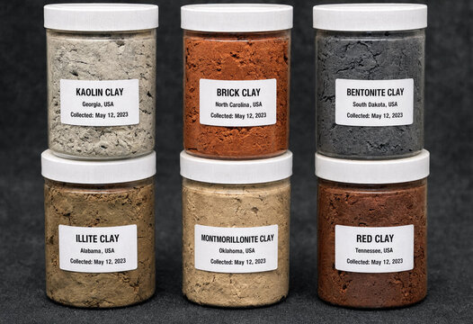 Six clear plastic jars containing different types of clay labeled with their names and collection locations on a dark grey background surface