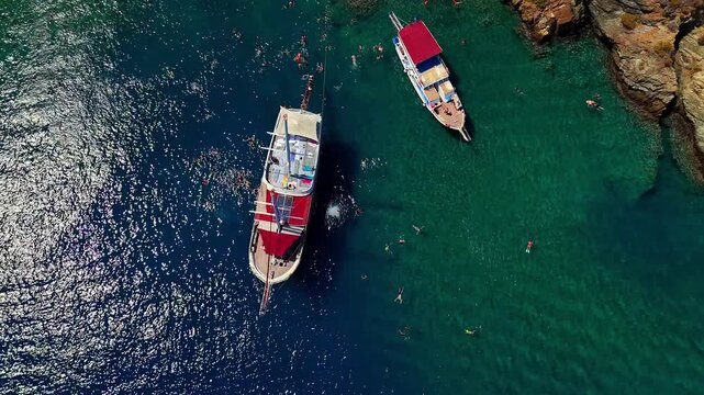 Aerial view of tour boats anchored in clear turquoise water with people swimming near a rocky coast, ideal for summer travel, tourism, vacation, cruise and Mediterranean lifestyle content.
