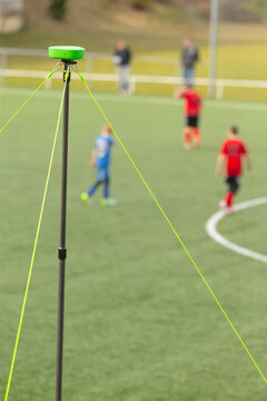 Tall surveying pole with a green top and neon guy lines set up on the sidelines of a turf soccer field. Out-of-focus youth players in red and blue kits are