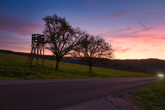 Hunting stand and trees by a country road under a colorful evening sky.