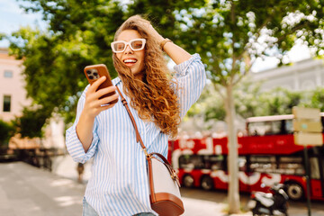 Fototapeta premium Portrait of a smiling woman with a phone relaxing on a bench on a sunny day. A beautiful woman with a phone enjoys time outdoors. Concepts: urban, leisure, and technology.