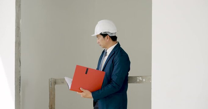 Asian male site manager walks with red document folder, inspecting unfinished wall surface, reviewing construction progress, building safety compliance, quality control, and contractor work plan.
