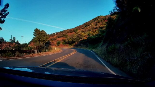 Vehicle travels on scenic mountain highway. Golden hour light illuminates rugged hills and roadside trees. Clear sky overhead, with light traffic on asphalt road. Dashcam perspective of journey.