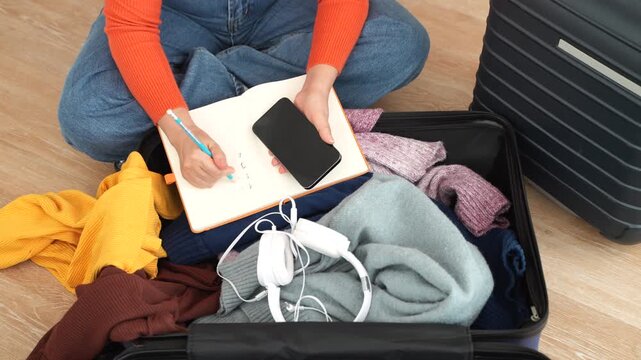 Hands writing a travel packing checklist in a notebook over an open packed suitcase, Concept of vacation preparation, trip planning, holiday organization, luggage management and journey readiness