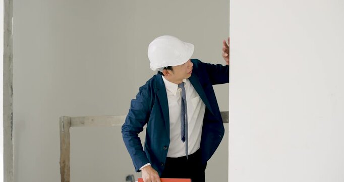 Asian female engineer wearing white helmet reviews orange folder, pointing toward unfinished wall during professional construction inspection, safety plan, and building progress assessment indoors.