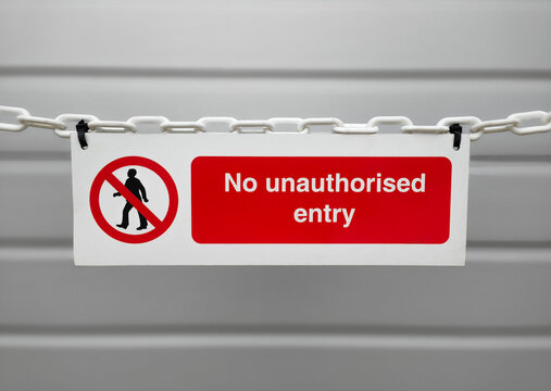 A red and white no unauthorised entry warning sign hanging from a white plastic chain, with a blurred grey industrial background. Concept of restricted access, security, safety, private area