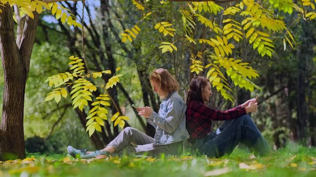 Two young women, a blonde and a brunette, are sitting back to back on a lawn among autumnal trees and using their smartphones. A loving LGBT couple enjoys a sunny autumn day in the park.