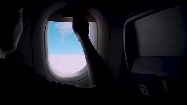 Silhouette hand lifting airplane window shade revealing bright sky during flight. Passenger reopening aircraft porthole cover inside dark cabin travel moment. Aviation interior contrast light and