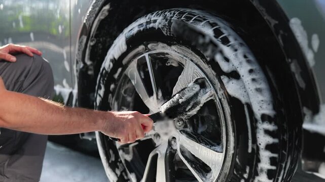 A person is meticulously cleaning a car's alloy wheel with a brush and soapy water