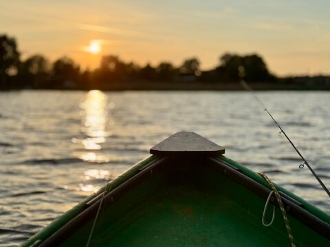 Point of view from a green boat on Liepāja Lake, Latvia, during a golden sunset. A fishing rod rests on the side with a calm water surface and silhouetted trees along the distant shore.