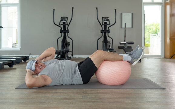 Senior chubby guy doing physical exercise with fitness ball lying on floor at gym. Side view of man having sport workout after gaining weight and getting fat.