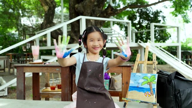 Cute happy Asian little girl showing her colorful painted hand, smiling child enjoying creative art workshop and beach canvas painting activity in the park