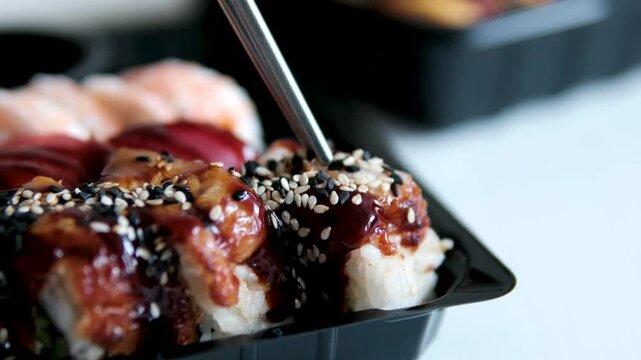 Metal chopsticks slowly glide through glossy unagi sauce and hover over an eel sushi roll sprinkled with white sesame. The moment captures anticipation, attention to flavor.