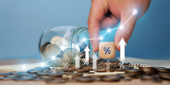 A close-up image shows a businessman's hand placing a percentage stick onto a pile of coins with an upward-pointing arrow on a graph. This image illustrates inflation management, interest rate hikes, 
