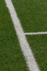 Fototapeta premium Shallow depth view of intersecting white field markings on dense green artificial turf. Visible texture of synthetic grass fibers, crisp paint edges and short