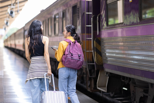 Two female tourists are walking on a train station platform towards a train, carrying luggage and backpacks, ready for their journey.