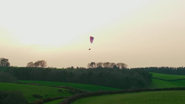 Paramotor pilot gliding above rolling green countryside hills and forest ridge during warm sunset sky. Powered paragliding drifting across rural landscape evening light extreme aerial adventure sport