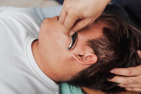 Therapist performing gua sha massage on a young male patient's jaw and neck area to relieve muscle tension and improve circulation.