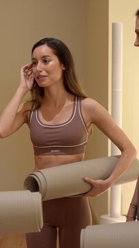 Two young athletic women in activewear holding yoga mats, smiling and talking together in a fitness studio before starting their workout, showing a healthy and active lifestyle
