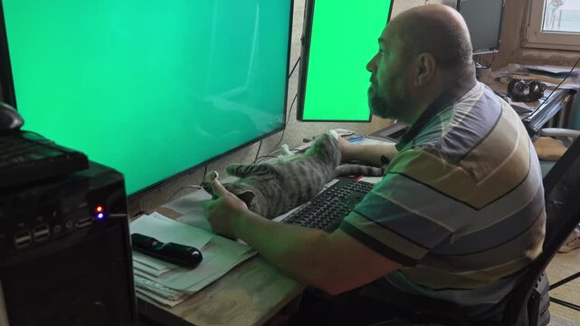 A middle-aged man handles business on dual computer monitors while his pet cat rests completely satisfied and stretched on the desk.