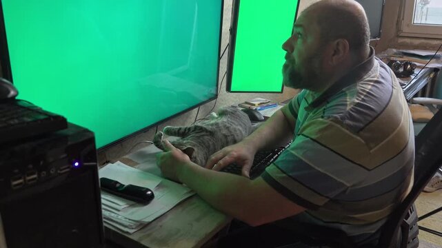 A middle-aged man handles business on dual computer monitors while his pet cat rests completely satisfied and stretched on the desk.