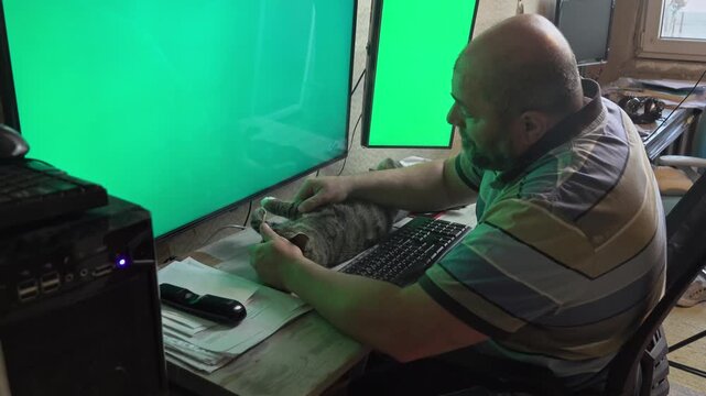 A middle-aged man handles business on dual computer monitors while his pet cat rests completely satisfied and stretched on the desk.