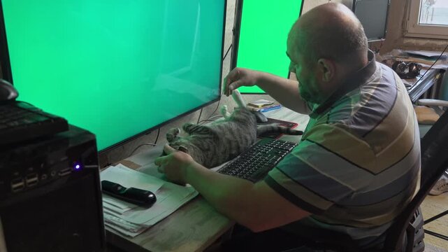 A middle-aged man handles business on dual computer monitors while his pet cat rests completely satisfied and stretched on the desk.