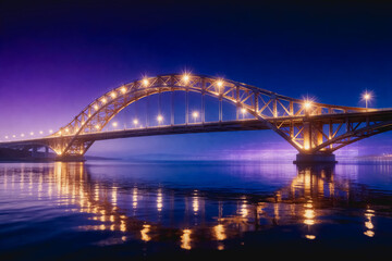Fototapeta premium An illuminated steel arch bridge spans calm water at dusk, with warm lights reflecting on the surface under a purple-blue sky.