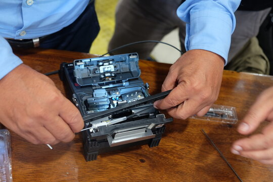 Technician hands splicing fiber optic cable using a professional fusion splicer machine on a table.