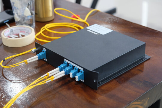 A black fiber optic patch panel with connected yellow cables on a wooden office desk surface.