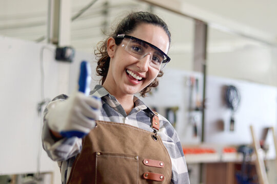 Portrait of happy smiling woman carpenter wears safety glasses goggles and apron, giving thump up, standing at carpentry workshop. Beautiful female craft worker creating wooden furniture at workplace.