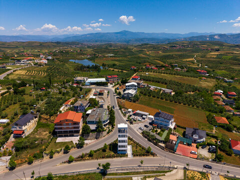 Aerial view of the village of Gradishte featuring houses with red roofs, a winding road, green fields, and a small lake under a clear blue sky in Gradishte, Fier County, Albania.