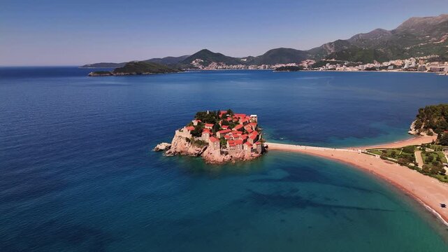 Sveti Stefan islet aerial view captures fortified island village with red tile roofs linked by sandy isthmus to Budva Riviera coastline amid clear Adriatic waters and mountainous backdrop for travel 