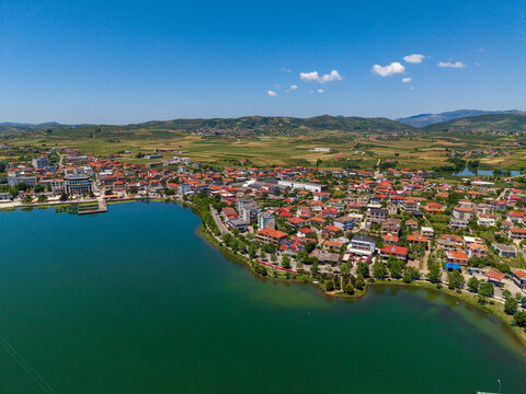 Aerial view of the town of Gradishte with red-roofed houses along the lakefront and rolling green hills under a clear blue sky in Gradishte, Fier County, Albania.
