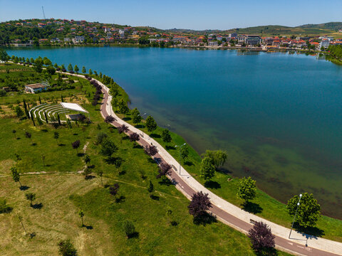 Aerial view of the waterfront promenade and green park area along the lake with a small amphitheater and town buildings under a clear blue sky in Gradishte, Fier County, Albania.