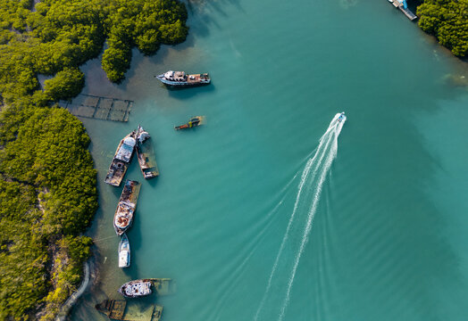 Aerial view of a boat leaving a white wake in turquoise water near rusted shipwrecks and lush green mangroves in Guanaja, Bay Islands Department, Honduras.