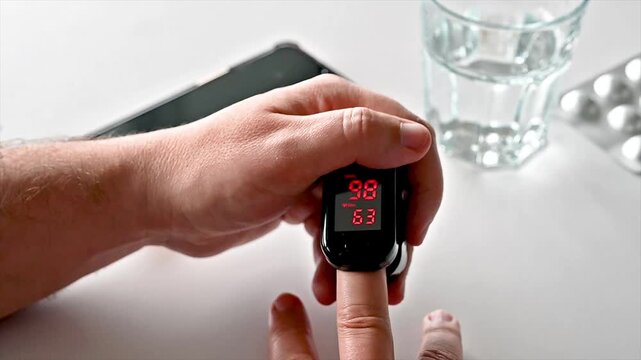 Close-up of a fingertip pulse oximeter displaying oxygen saturation on a mature man's finger at a table illustrating home health monitoring vitals checking and preventive self care.