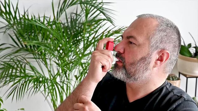 Close-up of a mature man taking a puff from a red asthma inhaler at home illustrating respiratory treatment breathing relief and chronic condition self management.