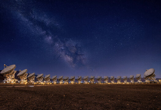 Large array of radio telescope satellite dishes in a desert landscape under a clear night sky filled with the bright stars of the milky way