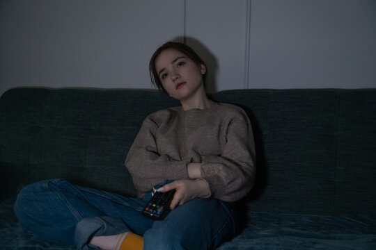 Girl sitting on a couch in a dark room at night, holding a remote control and feeling bored, showing concepts of loneliness, relaxation, and staying at home