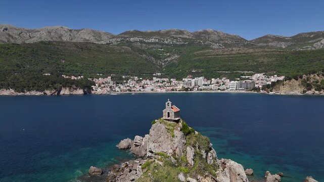 St Nedjelja Church sits on rugged islet off Petrovac coast captured by drone, showcasing turquoise Adriatic waters, rocky promontory, coastal town and verdant hinterland mountains, drone shot