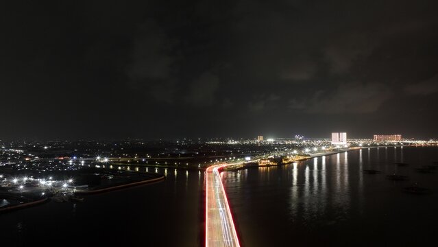 Aerial view of the PIK 2 bridge at night with long exposure traffic light trails over the water towards the city skyline in Kecamatan Cakung, Daerah Khusus Ibukota, Indonesia.