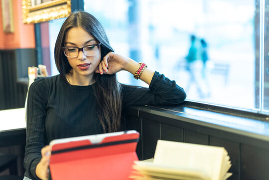 Pretty, brunette woman has a coffee in a bar while she looks at her tablet and consults a book