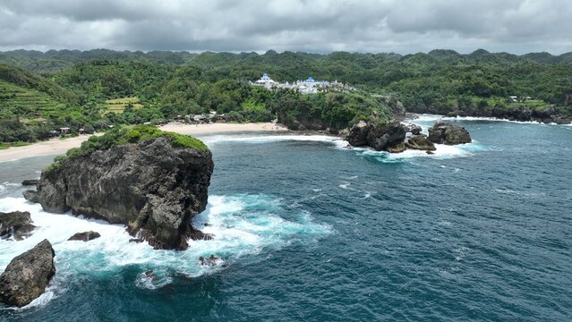 Aerial view of Jungwok Beach featuring massive rock formations, crashing waves, and lush green hills in Girisubo, Special Region of Yogyakarta, Indonesia.
