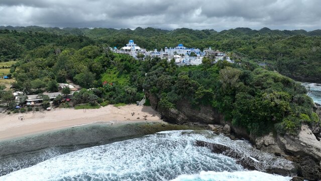 Aerial view of Jungwok Beach with white and blue buildings on a lush green cliff overlooking the sandy shore and ocean waves, Girisubo, Special Region of Yogyakarta, Indonesia.
