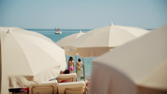Cinematic summer beach scene with tourists relaxing on sun loungers beneath large white umbrellas by the sea