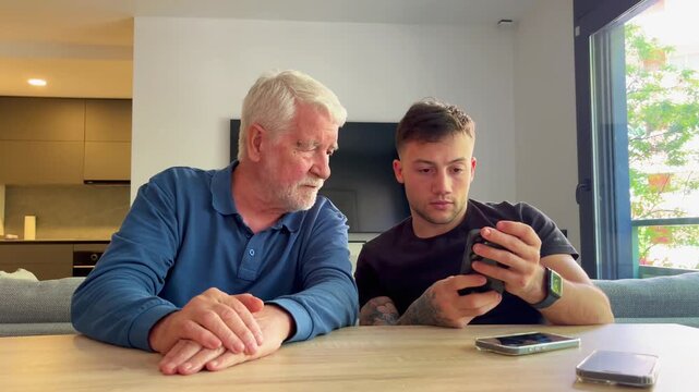 Young man explaining how a mobile phone works to his curious elderly grandfather, sharing knowledge between generations and bridging the digital divide with patience and modern technology at home