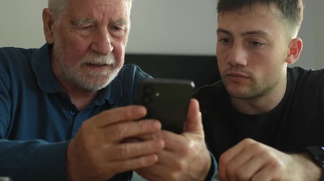 Two generations, a grandfather and his grandson, are together looking at a smartphone screen, with the younger man patiently explaining how the modern device works to the senior man